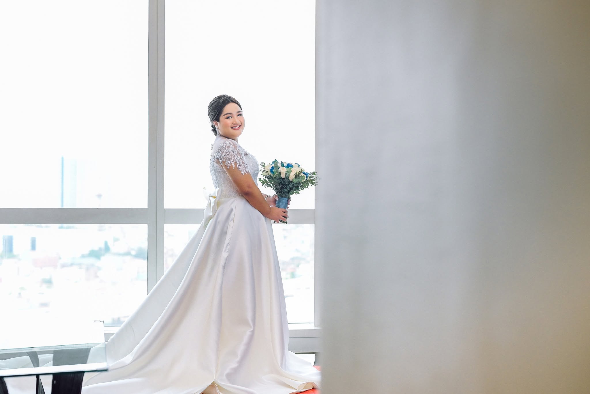 Bride in a white gown holding a blue and white bouquet by a bright window